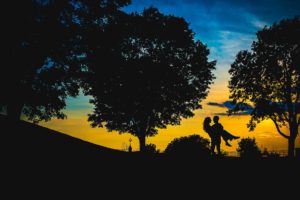 silhouette of groom holding bride in front of big blue and orange sky